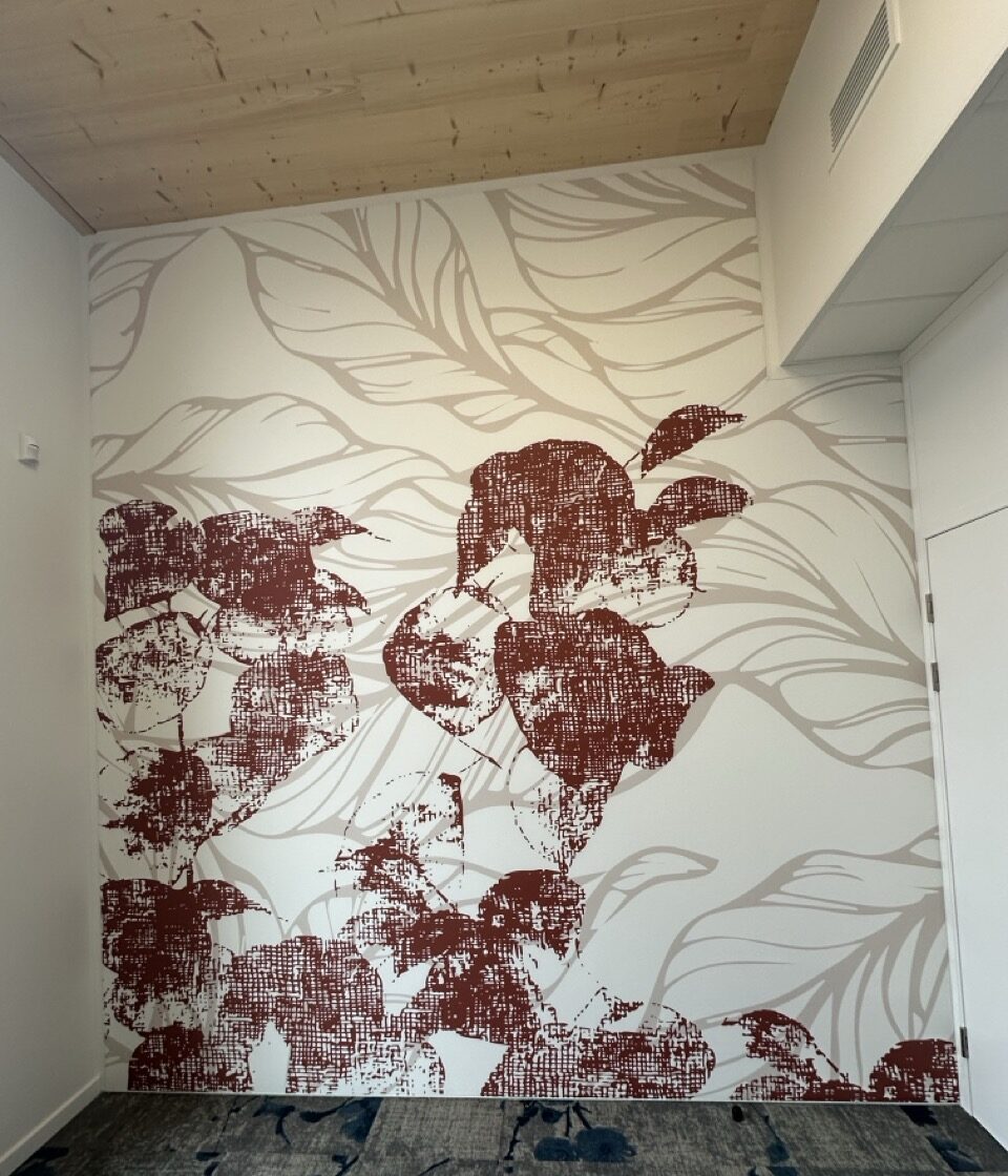 Angle de pièce habillé d’une toile tendue imprimée d’un motif de feuillages, feuilles beiges dessinées au trait et feuilles terracotta texturées, sous un plafond en lambris clair