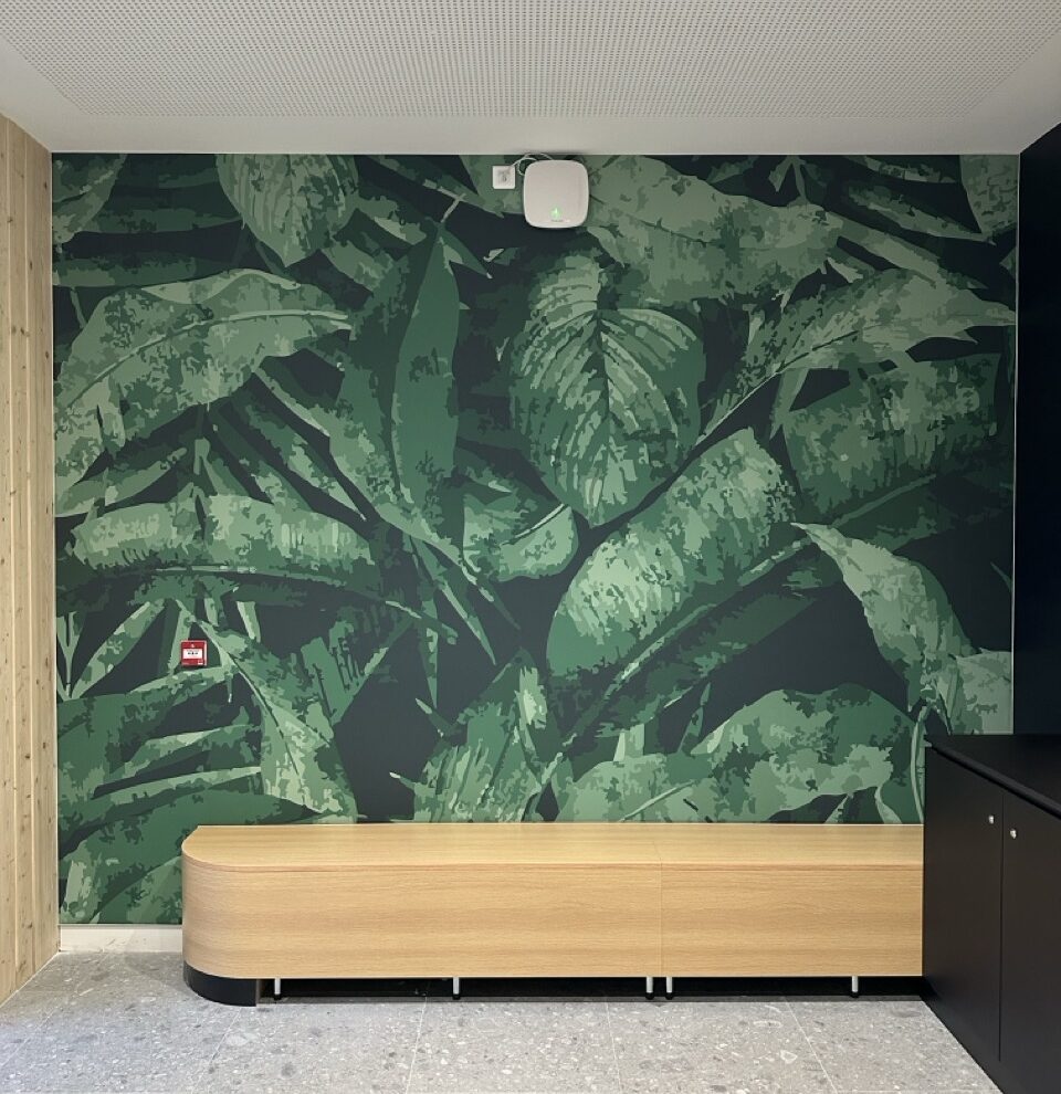 Mur d’un hall d’accueil entièrement couvert d’une toile tendue imprimée d’un motif de feuillages verts, devant une banque d’accueil en bois clair et un sol en terrazzo