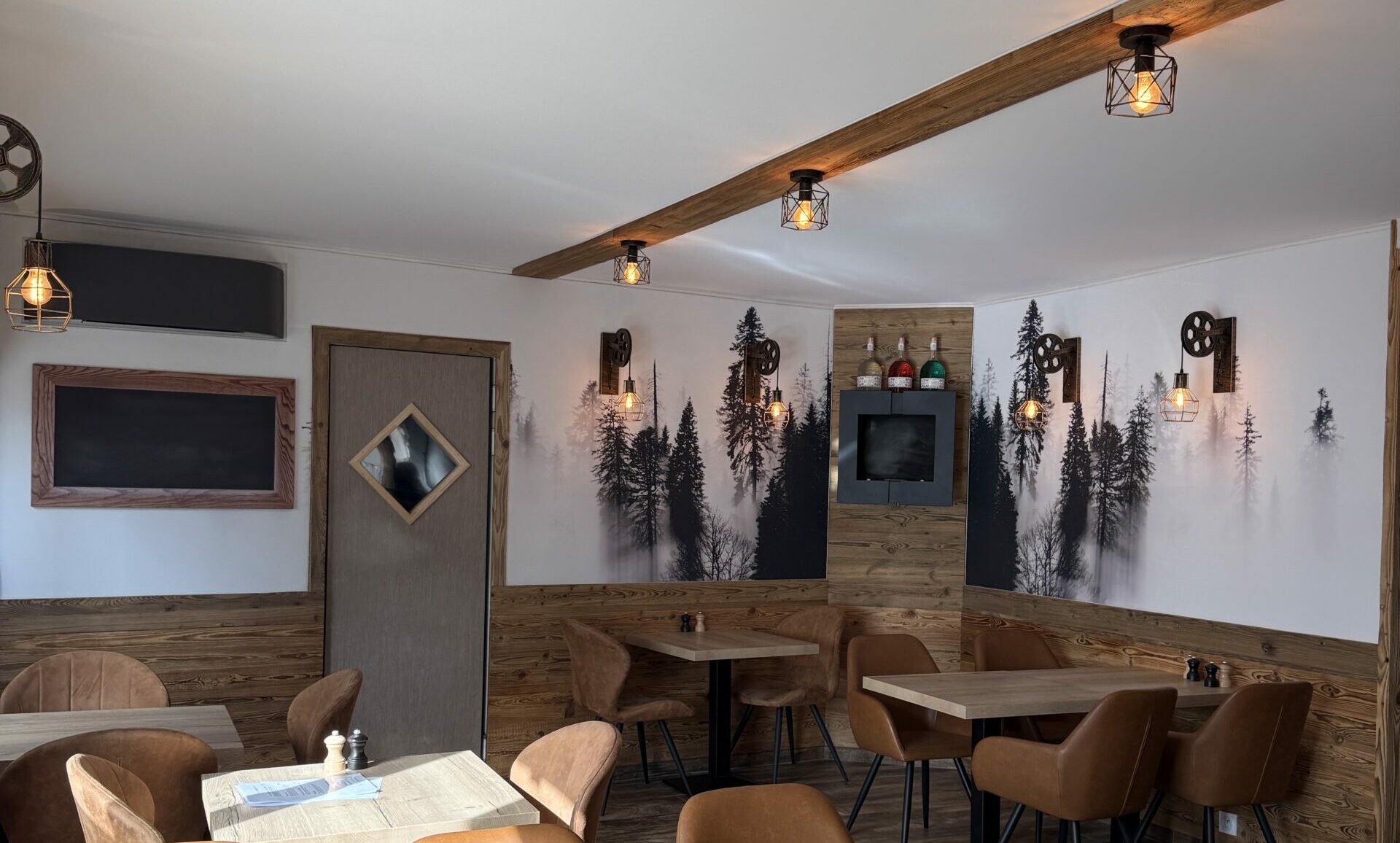 Salle de restaurant dont les murs portent une toile tendue imprimée d’une forêt de sapins dans la brume, au-dessus d’un lambris de vieux bois, sous un plafond tendu blanc