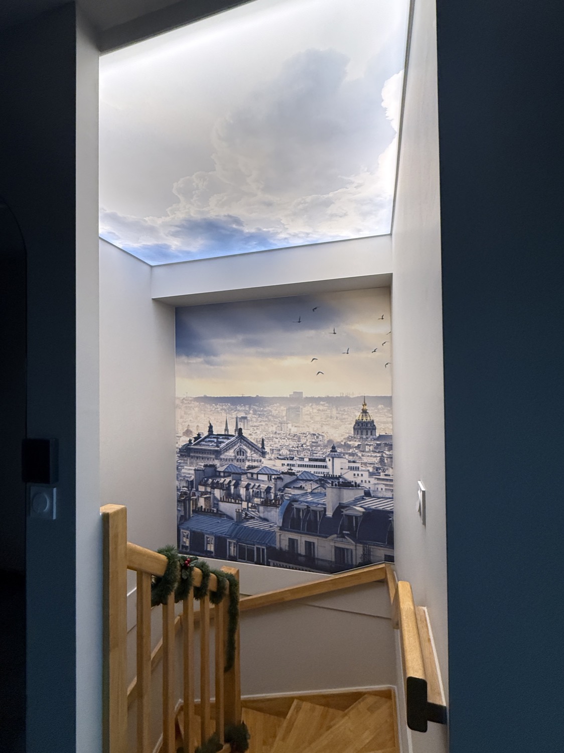 La même cage d’escalier après intervention : le mur du fond porte une toile tendue imprimée des toits de Paris, et le plafond de la trémie une toile translucide rétroéclairée imprimée d’un ciel nuageux
