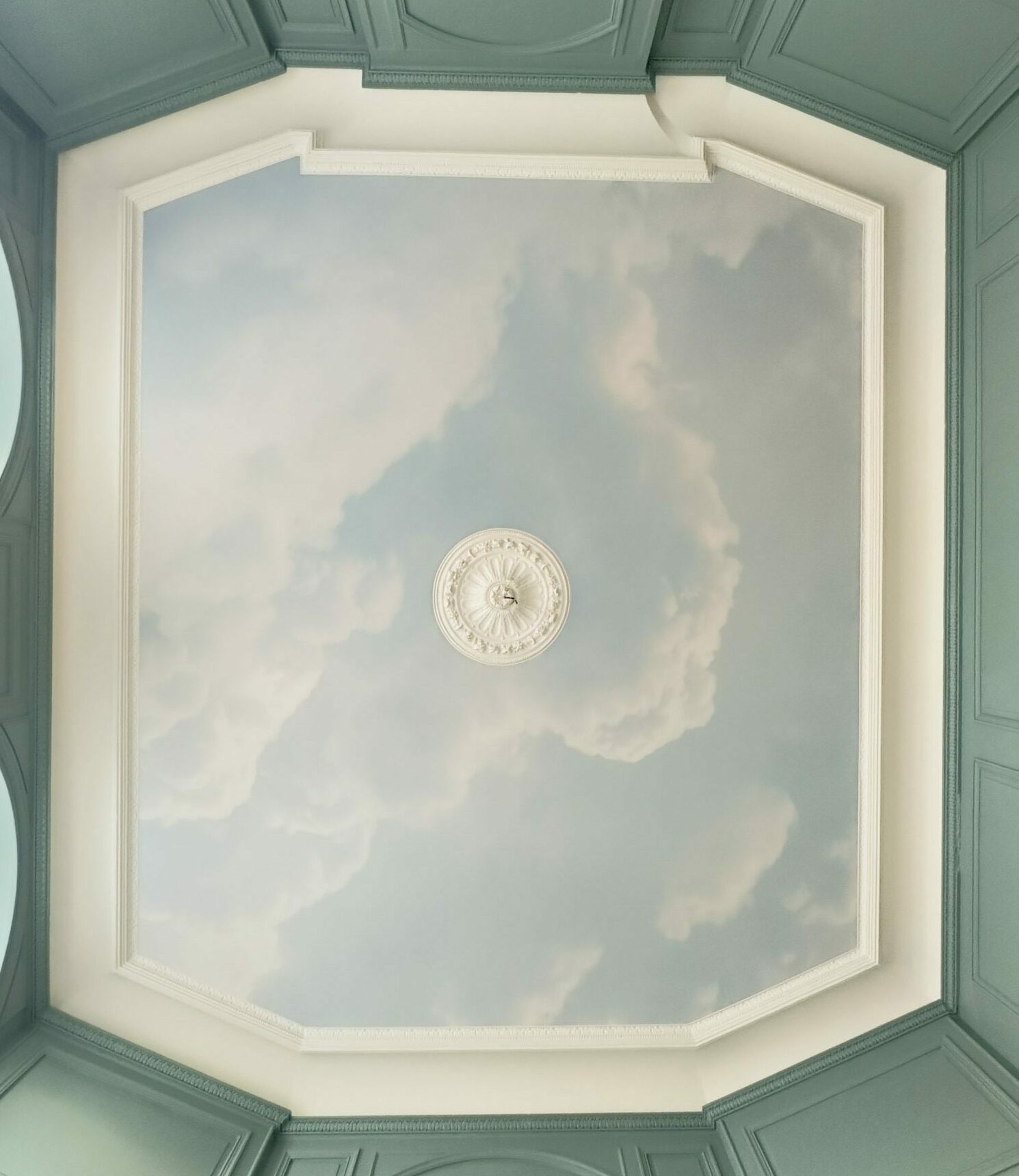 Plafond tendu imprimé d’un ciel nuageux dans un caisson octogonal à moulures blanches, entouré de boiseries vert amande, avec une rosace de plâtre au centre