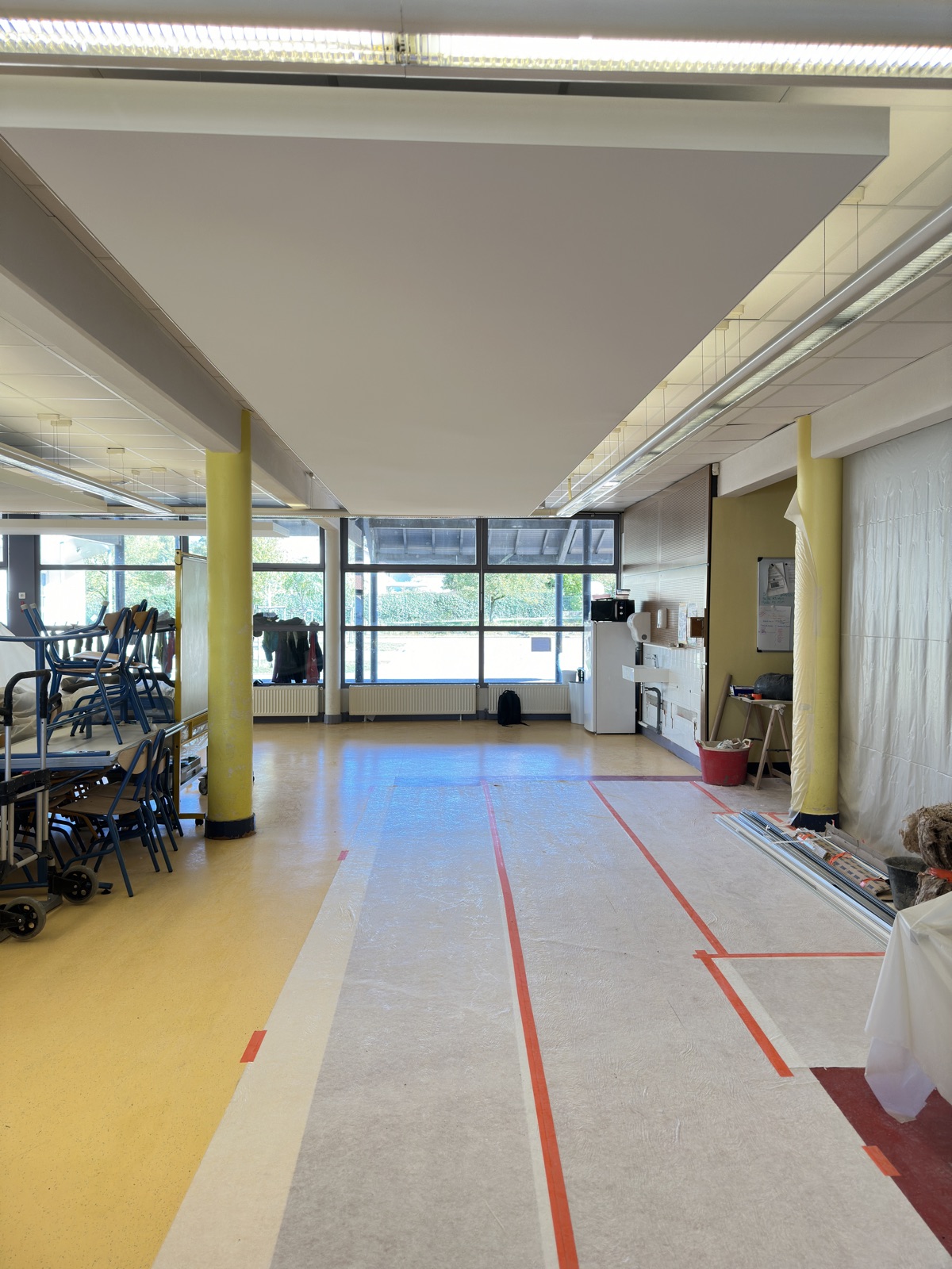 Grand îlot acoustique blanc suspendu au-dessus d’une salle de restauration scolaire, chaises empilées sur le côté