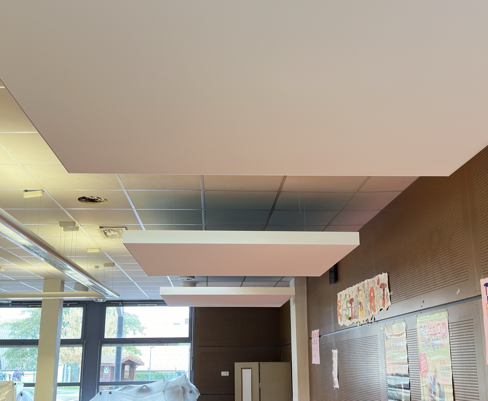 Trois îlots acoustiques blancs suspendus sous le plafond à dalles d’un réfectoire d’école